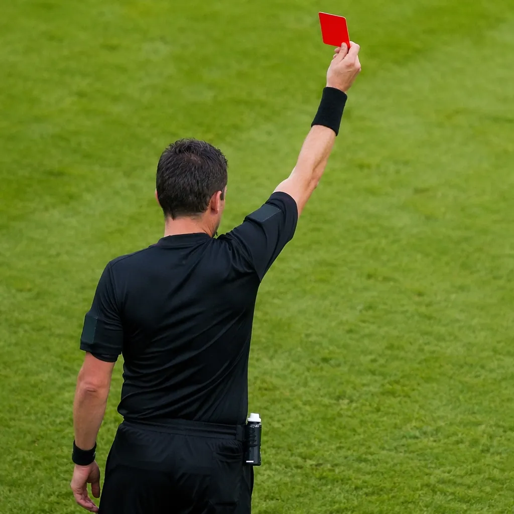 Photo of a football referee holding a red card on a green pitch, no identifiable