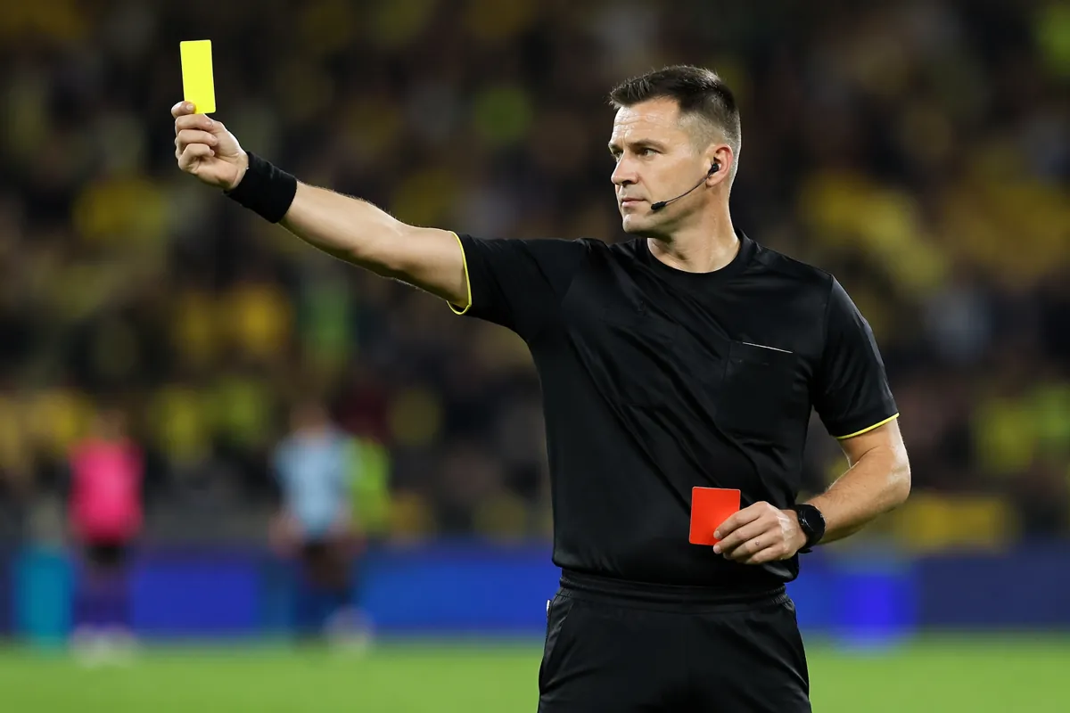 Professional football referee holding a yellow and red card on a green football pitch, bri