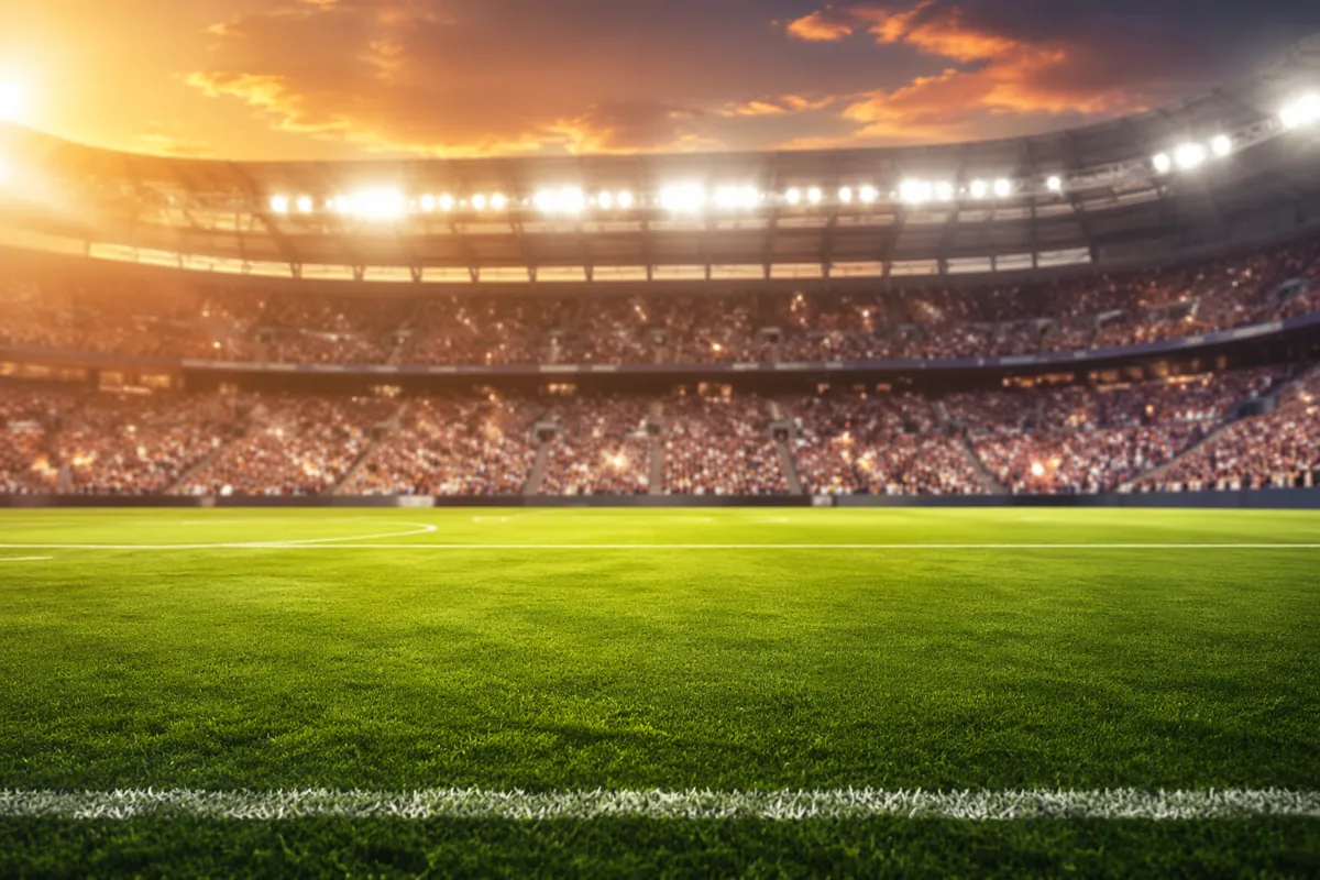 Wide shot of a green football pitch under bright stadium lights, blurry cheering fans in t