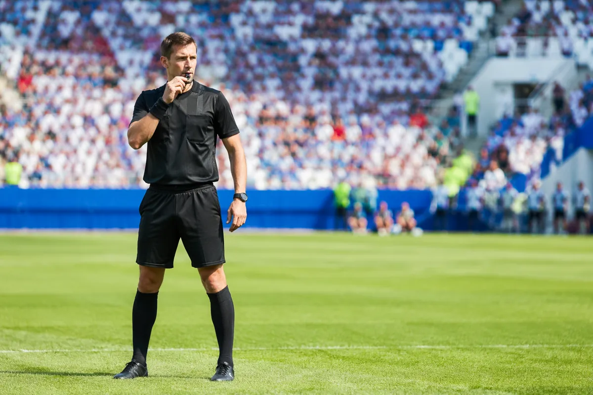 Wide shot of a professional soccer referee holding a whistle, standing on a green soccer f