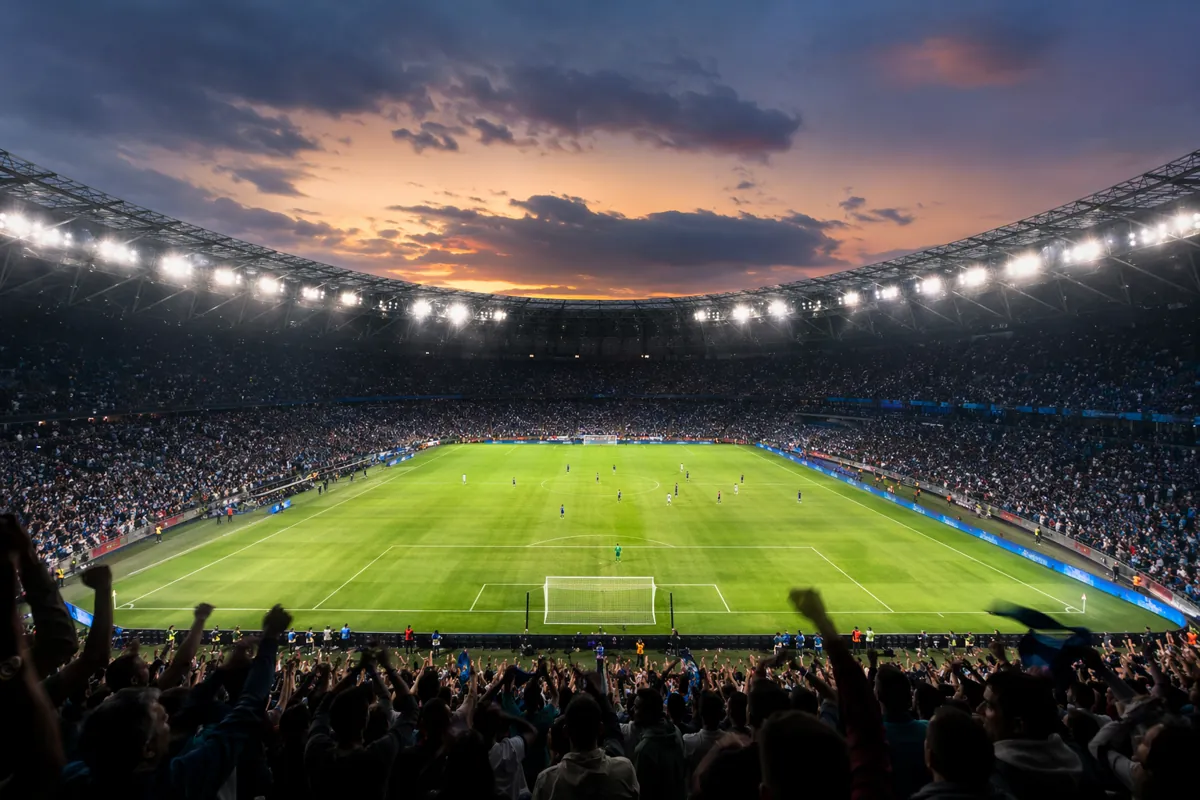 Wide shot of a generic football stadium at dusk, with green pitch, glowing stadium lights,