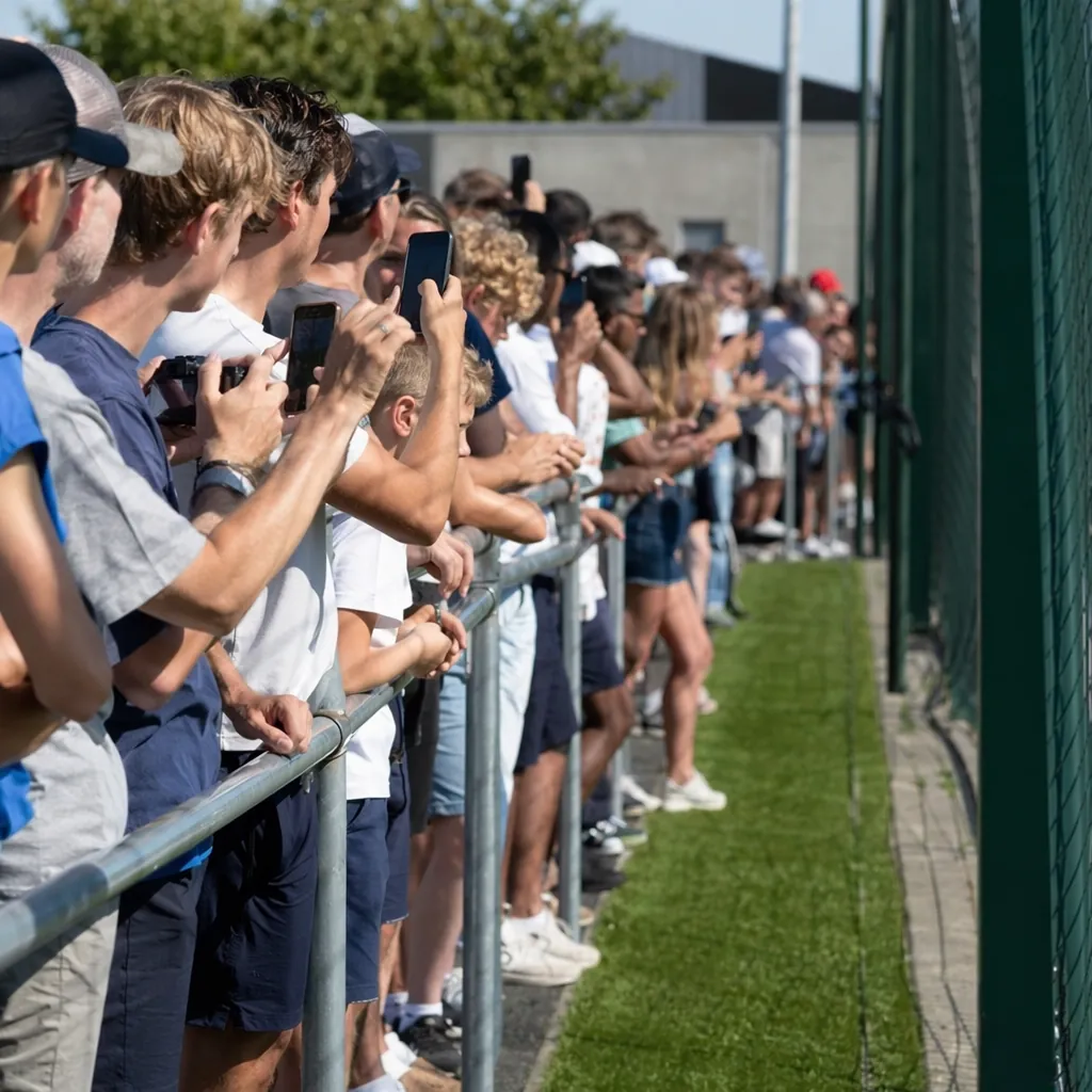 Side view of fans standing behind barrier outside football training ground, hold