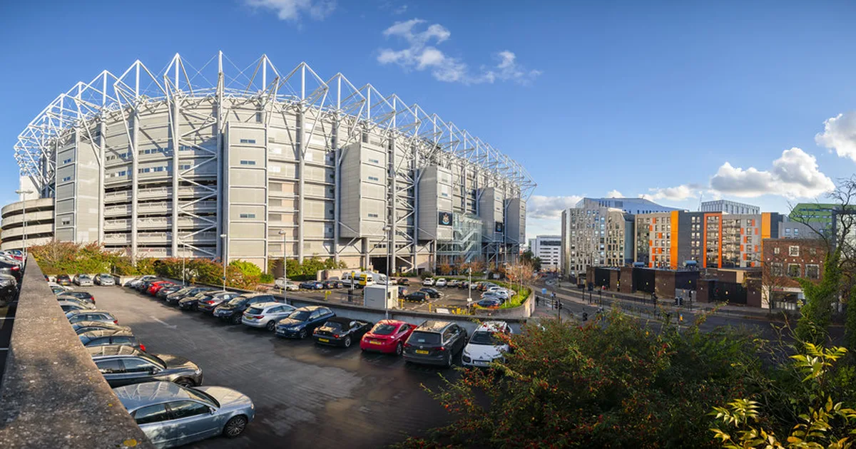 Wide shot of a modern football stadium at dusk, empty green pitch in the foreground, blurr