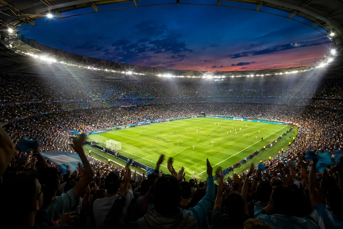 Wide shot of a packed world cup football stadium at dusk, bright floodlights illuminating