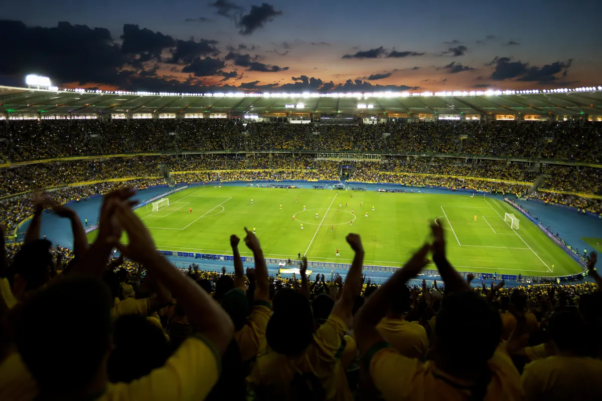 A professional football stadium at dusk, bright floodlights illuminating the green pitch,