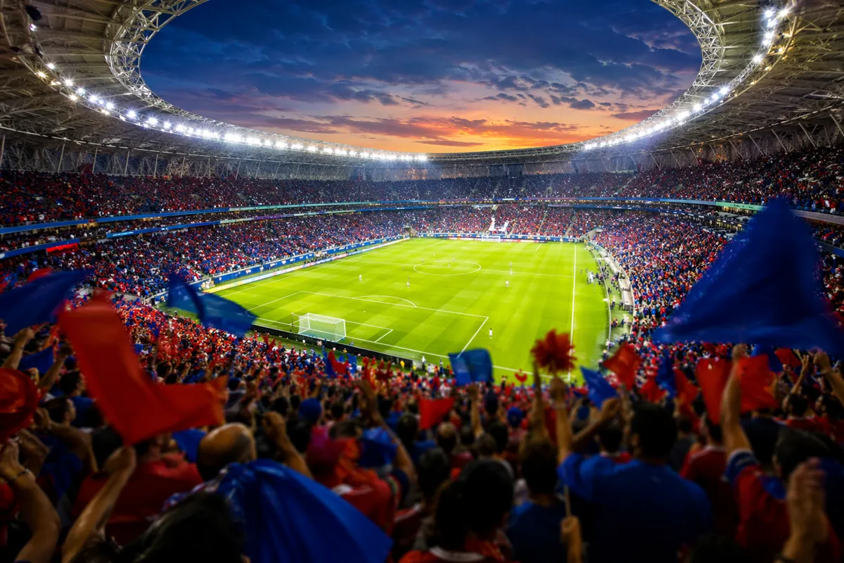 Wide shot of a packed modern football stadium at dusk, vibrant green pitch, blurred crowd