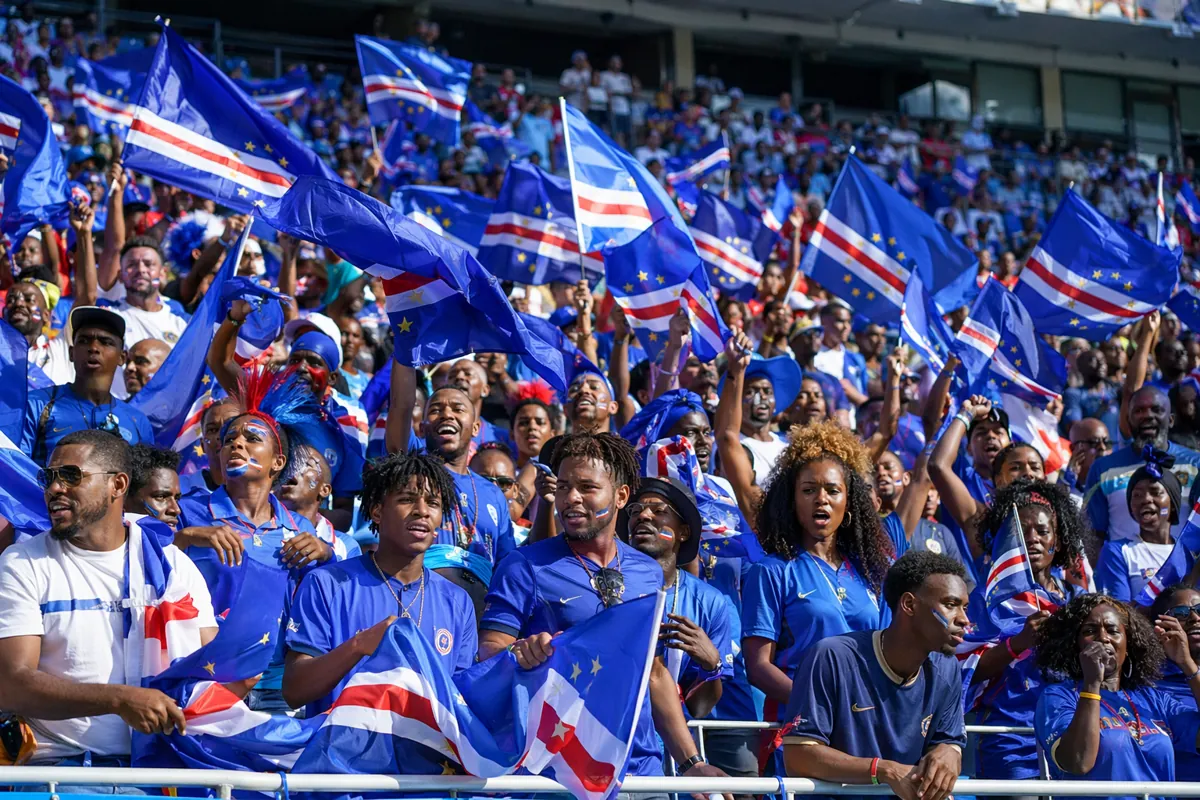 Wide shot of football fans waving Cape Verde national flags in a stadium, vibrant match at